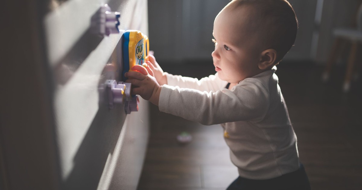 A cute baby exploring magnetic toys on a cupboard, illuminated by natural light indoors.