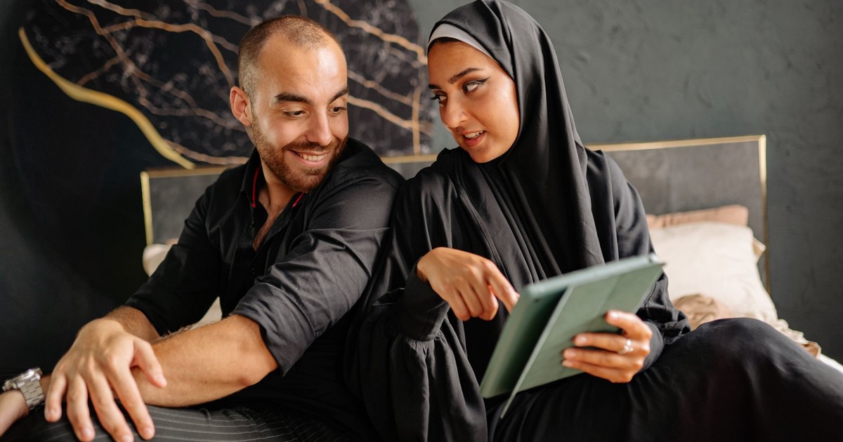 A diverse couple sitting on a bed and discussing something on a tablet indoors.