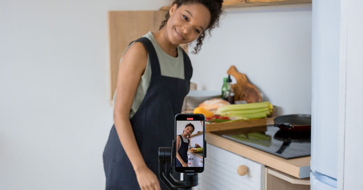 Smiling young woman films a cooking video at home using a smartphone in her kitchen.