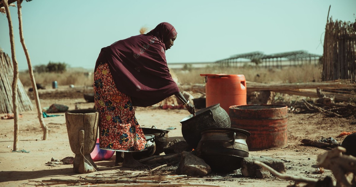 African woman cooks outdoors in a traditional setting with pots over an open fire in a desert landscape.