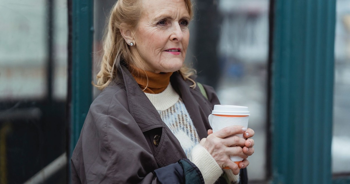Contemplative elderly female in outerwear with hot beverage to go looking forward in town in daylight