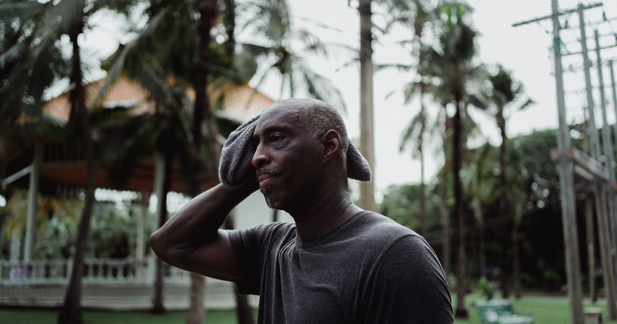 A man wiping sweat with a face towel outdoors in a tropical environment, appearing thoughtful and serious.