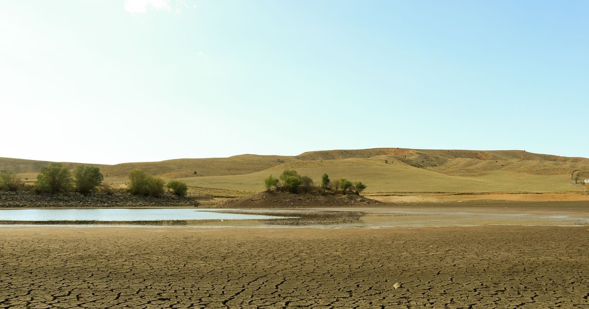 A vast arid landscape showing cracked earth, water, and sparse greenery under a clear sky.