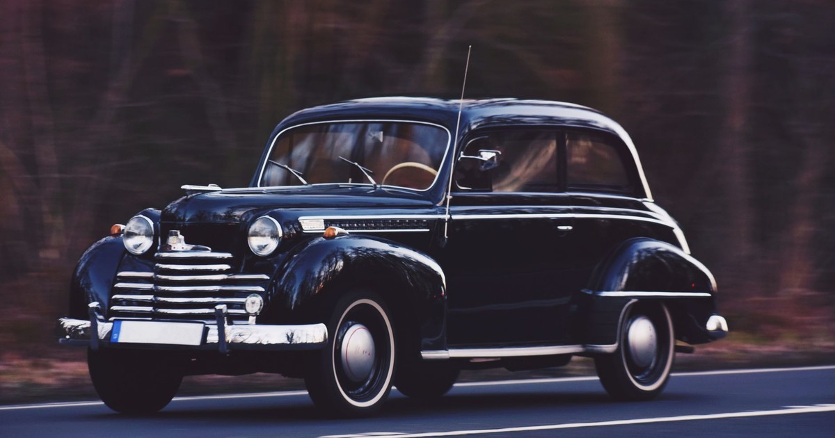 A sleek vintage car driving on a road with blurred wooded background, showcasing timeless elegance.
