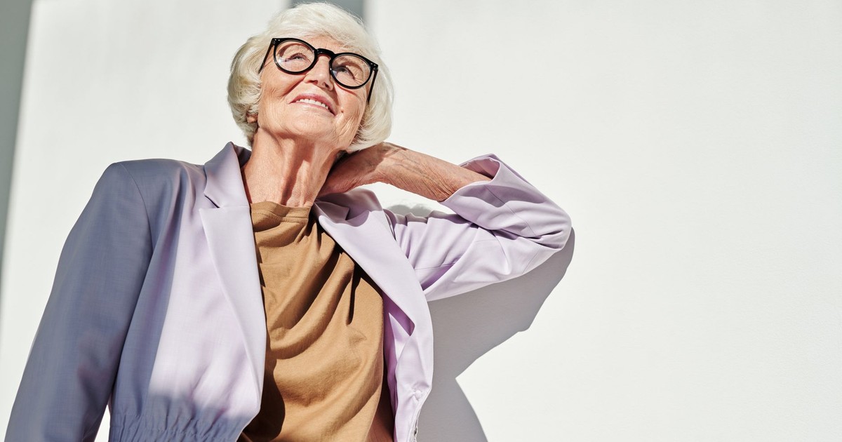 A senior woman wearing fashionable eyewear and attire smiles confidently in bright daylight.