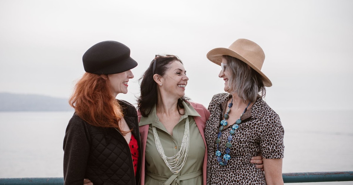 Three women in stylish outfits smiling together outdoors by the sea.