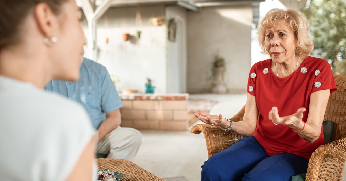 Senior woman in a red shirt chatting expressively with another woman in a cozy setting.
