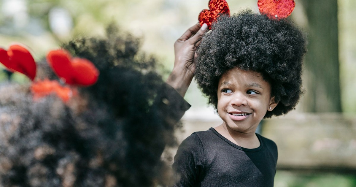 A girl with afro hair smiles as her parent places heart-shaped accessories on her head outdoors.