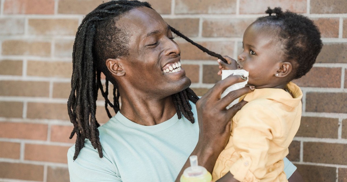 Smiling father with dreadlocks lovingly engages with his baby daughter outdoors against a brick wall.