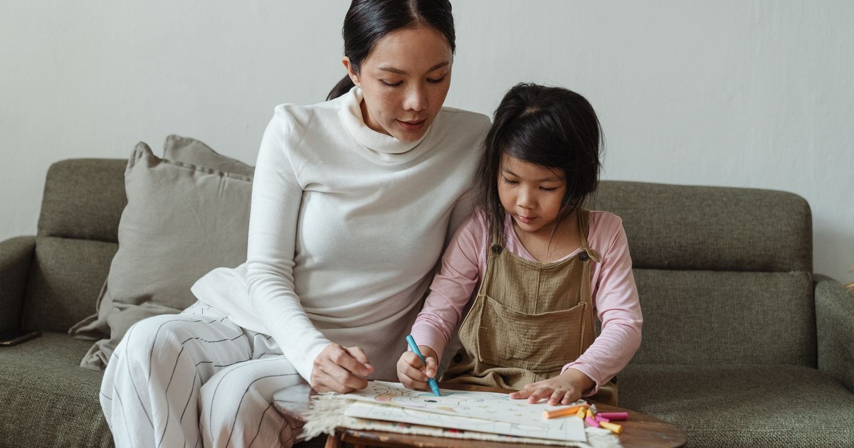 Concentrated little Asian girl in casual clothes studiously drawing picture on paper while mother watching process sitting together on sofa