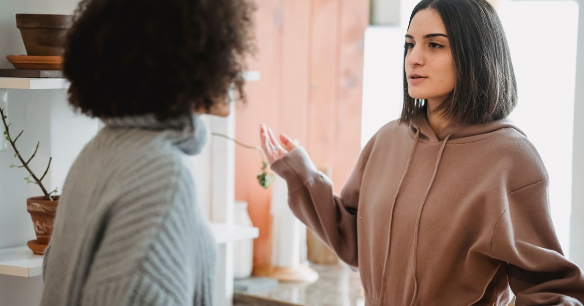 Serious woman in casual wear having argument with anonymous female with curly hair while standing in light room at home