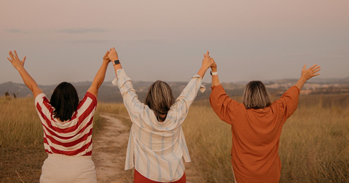 Three women standing in a São Paulo field, holding hands and raising arms, celebrating friendship.
