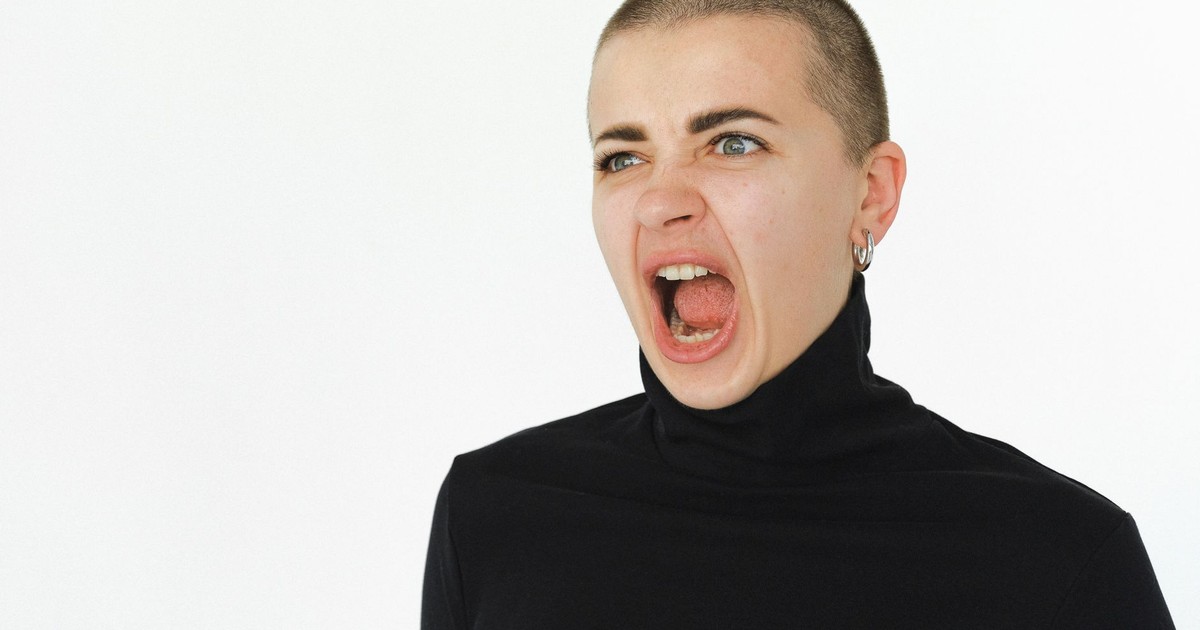 A woman yells expressively in a studio setting, wearing a black turtleneck.