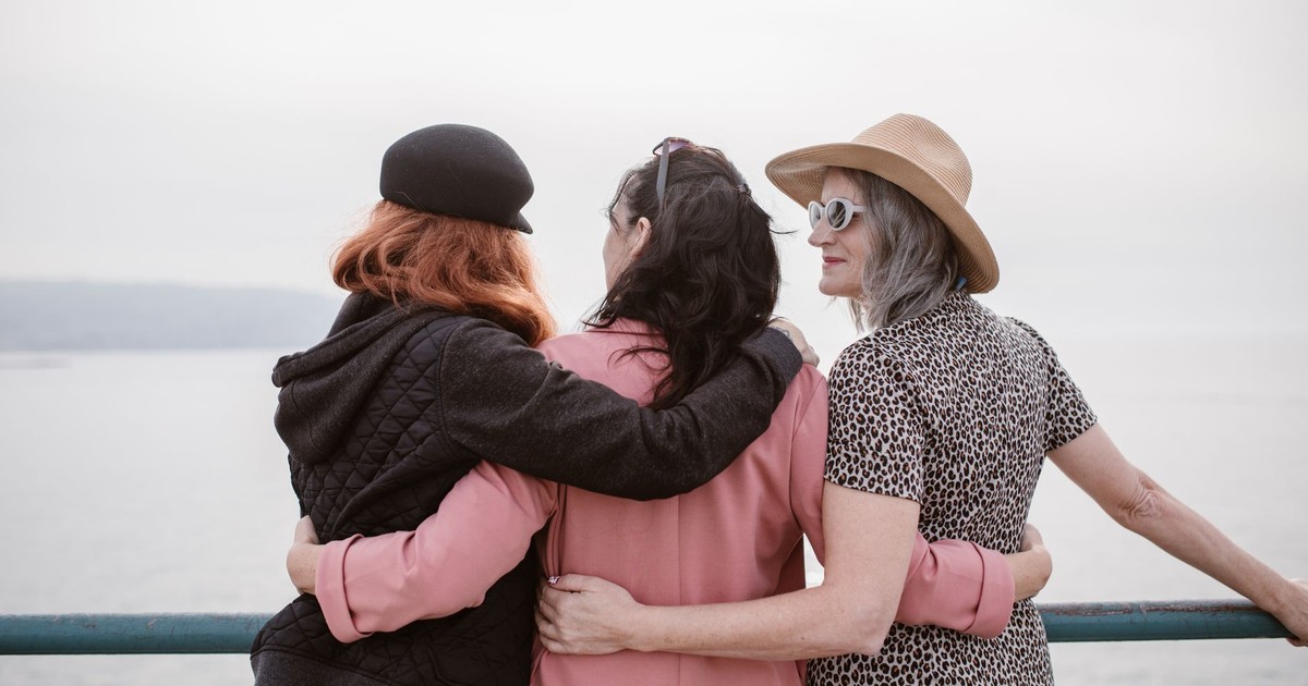 Three women, seen from behind, embracing by a seaside railing, symbolizing friendship.