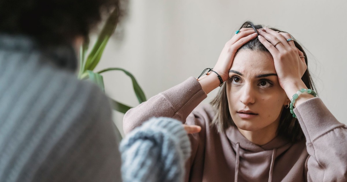 Worried female sitting near wall with hands on head and looking at faceless female while having conflict in light room