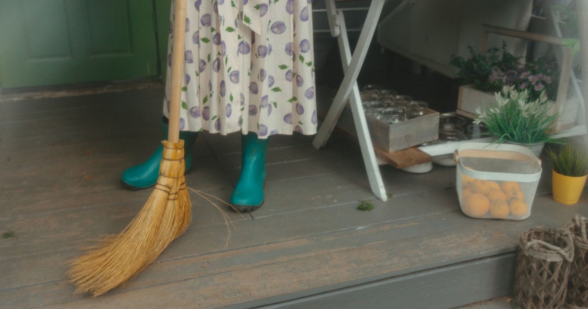 A woman in a floral dress and rubber boots sweeps a wooden porch beside a table with plants.