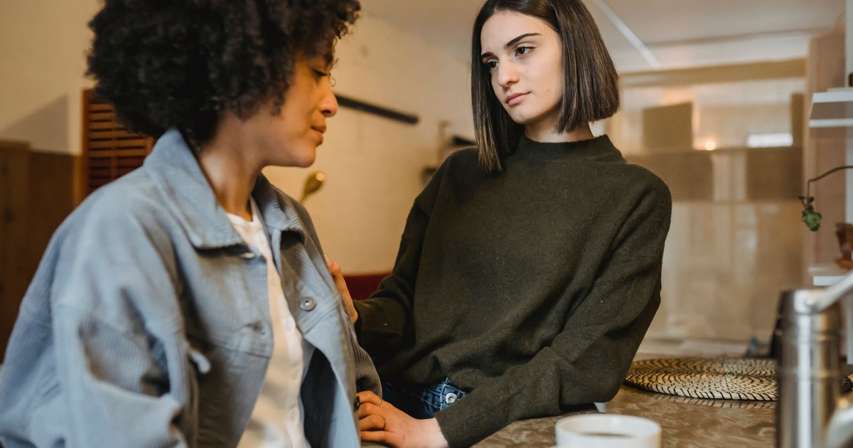 Delicate female holding hand of upset African American female having problem sitting at counter with cup of coffee