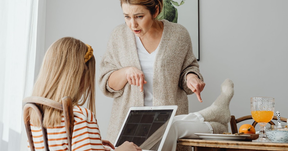 A mother scolds her teenage daughter using a laptop at home, highlighting a generational clash.