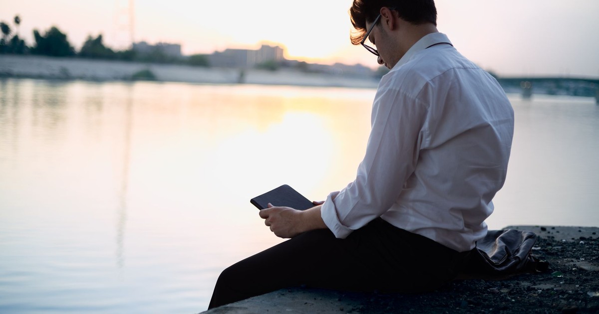 A businessman seated by a riverbank, reading on a tablet as the sun sets, creating a peaceful and reflective scene.